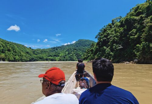 Four people, one wearing a red baseball cap and another a blue shirt, ride a boat on a khaki-colored river surrounded by dense forest.