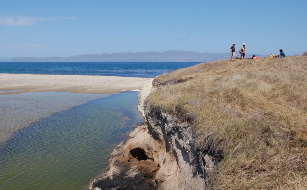 Several people stand on a grassy cliff with green shallow water off to their left and blue water and a mountain range in the distance.