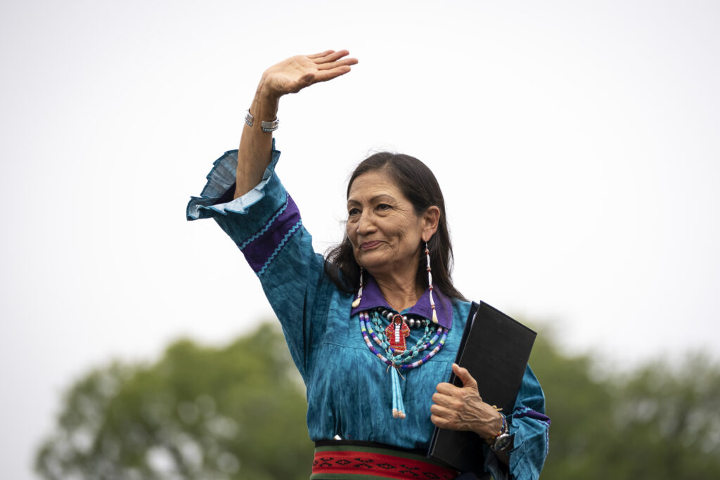 A smiling person with long, straight black hair wearing a teal and purple top with embroidered details, red patterned waistbelt, and beaded necklace and earrings holds a black folder in their left hand and waves with their right hand.