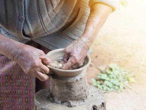 A close-up image features the wrinkled hands of a person wearing a cardigan and printed skirt as they mold clay into a bowl shape.