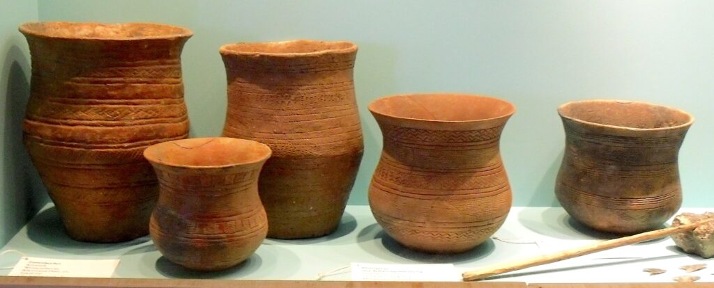 Small tan pots that narrow in the middle and flare out at the top sit in a row on a long white shelf behind two small pieces of paper with text printed on them.