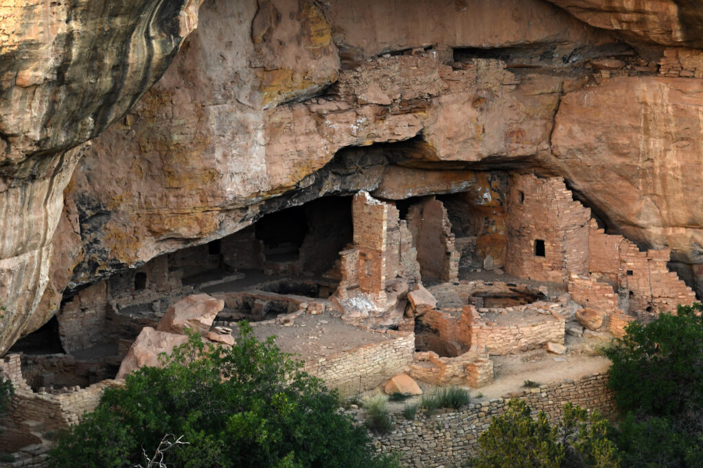 From far away, a landscape of circular sandstone structures sits to the left of a sandstone wall with a few rectangular windows. All structures are built into a large opening on the side of a desert cliff wall.