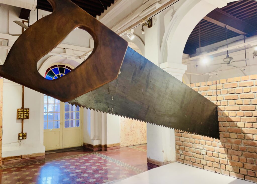 Inside a room with a white arch and red, white, and blue tiled floor, a large saw sits in a tan brick wall, appearing to have cut it.