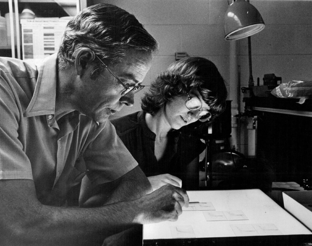 A black-and-white photograph features two people wearing glasses seated in an office in front of a light table. Both look down at microscope slides on the table.