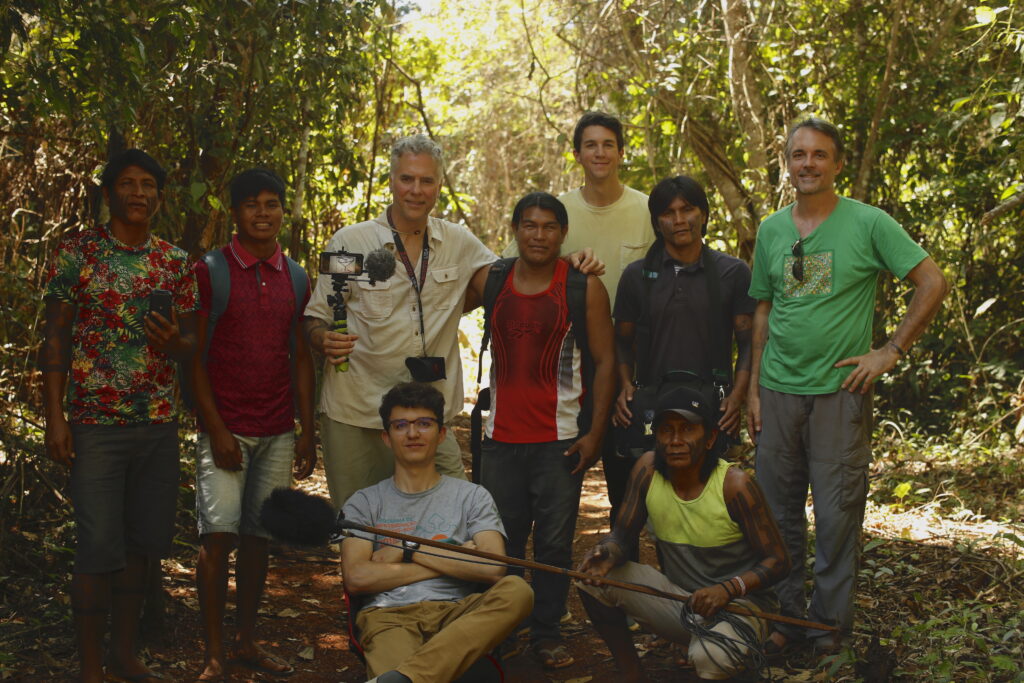 A group of nine people—one holding a camera and another holding a microphone on a long wooden stick—pose in a leaf-covered opening in a forest.