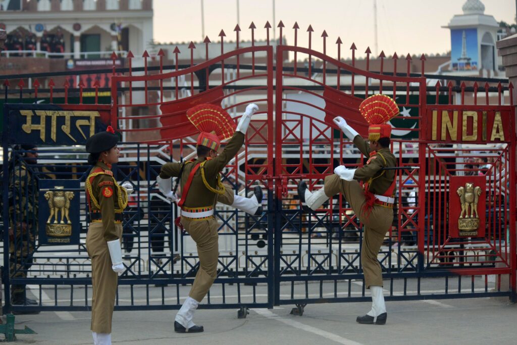 People wearing matching uniforms of green jackets, khaki pants, and black boots stand in front of a red and black gate that has the word “India” on it in gold letters. Two people put their legs on the gate to help push it shut.