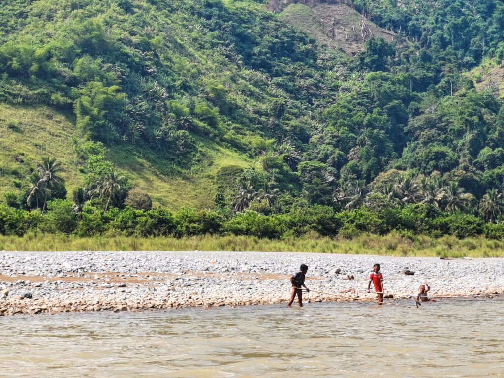 Duas crianças seguram bastões de madeira enquanto estão em um rio, e uma terceira criança senta-se no rio em frente a uma margem rochosa e a uma montanha coberta pela floresta.