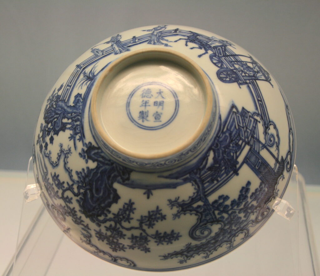 An overturned, ornately decorated blue-and-white bowl is on display on a plastic stand, revealing Chinese characters on its bottom.