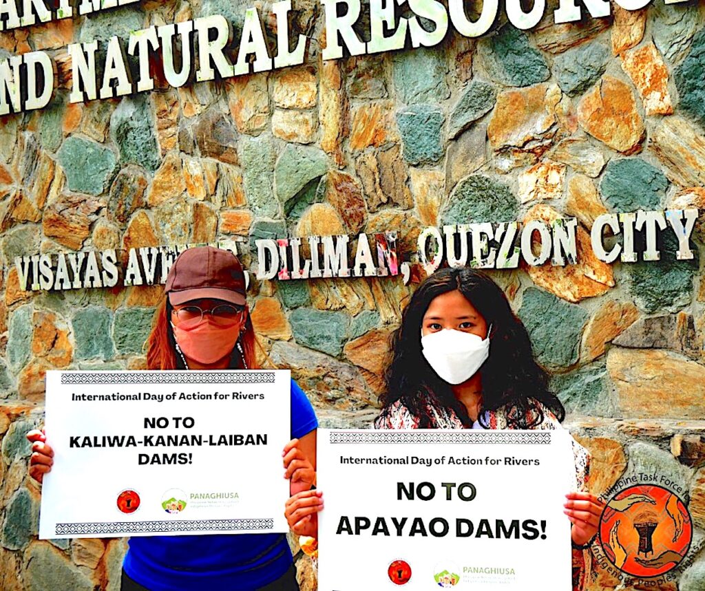 Duas pessoas usando máscaras seguram cartazes que dizem “Não às represas de Kaliwa-Kanan-Laiban” e “Não às represas de Apayao”.