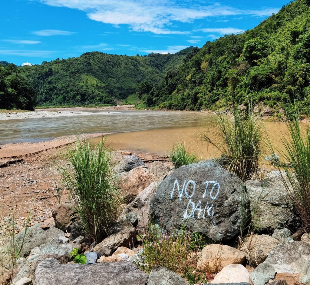 Um rio corre ao longo de uma margem onde há uma pedra pintada com as palavras “Não à barragem".