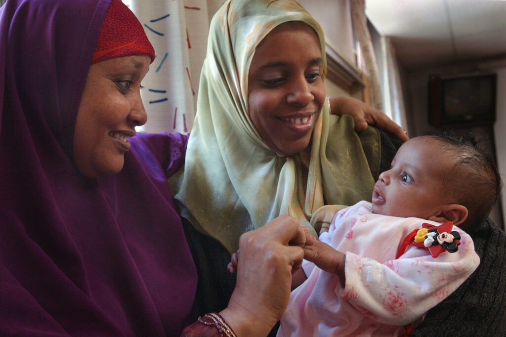Two people who are wearing purple and olive-green hijabs, respectively, are holding and gazing at a baby in a pink onesie.