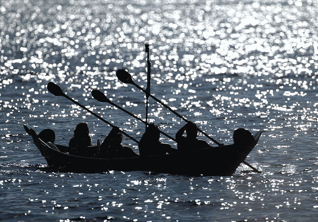 A photograph features the black silhouette of several people rowing a canoe against a background of grayish-blue water that has light reflecting off its surface.