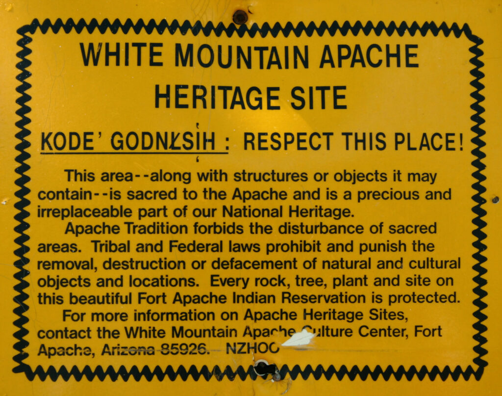 A yellow sign has a squiggly black border around a block of text. The header reads, in all capital letters, “White Mountain Apache Heritage Site.” Below this are underlined words in the Ndee language translated to “Respect This Place” in English followed by an exclamation point. Below this, a paragraph reads, “This area—along with structures or objects it may contain—is sacred to the Apache and is a precious and irreplaceable part of our National Heritage. Apache Tradition forbids the disturbance of sacred areas. Tribal and Federal laws prohibit and punish the removal, destruction, or defacement of natural and cultural objects and locations. Every rock, tree, plant, and site on this beautiful Fort Apache Indian Reservation is protected. For more information on Apache Heritage Sites, contact the White Mountain Apache Culture Center, Fort Apache, Arizona 85926. NZHOO.”