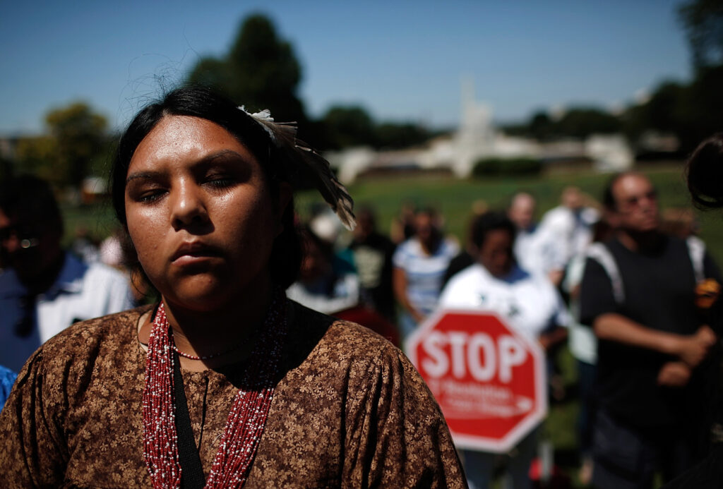 A person wearing a brown floral shirt, red beaded necklace, and feather in their hair closes their eyes. In a blurred background, other people—one holding a red-and-white STOP sign—gather on a lawn in front of a white building.