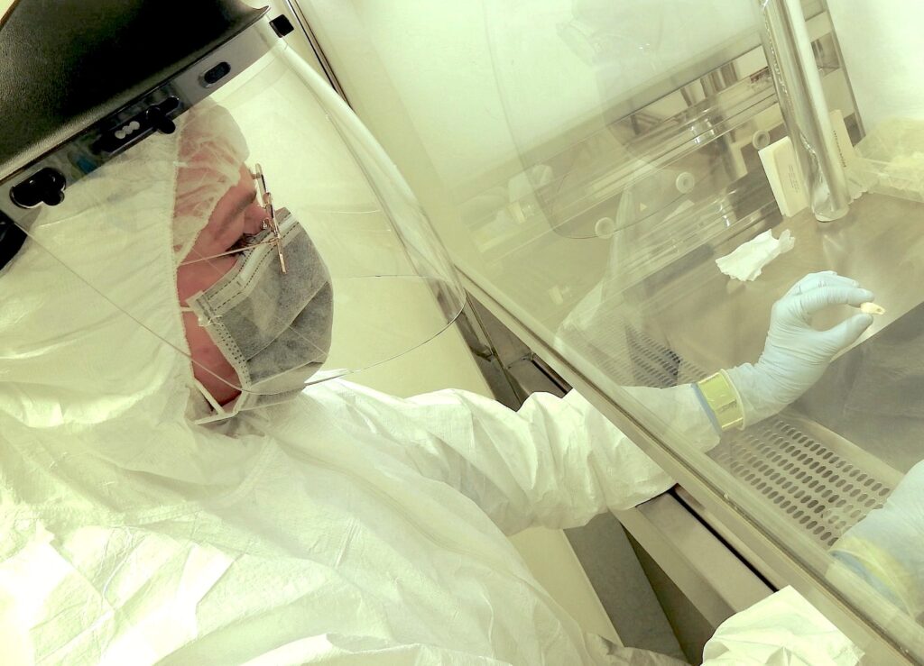In a science lab, a masked person wearing a white hazmat suit, face shield, and bouffant cap holds small objects under the glass hood of a silver metal workstation.