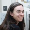 A woman with long brown hair turns to face the camera in front of a scientific instrument.