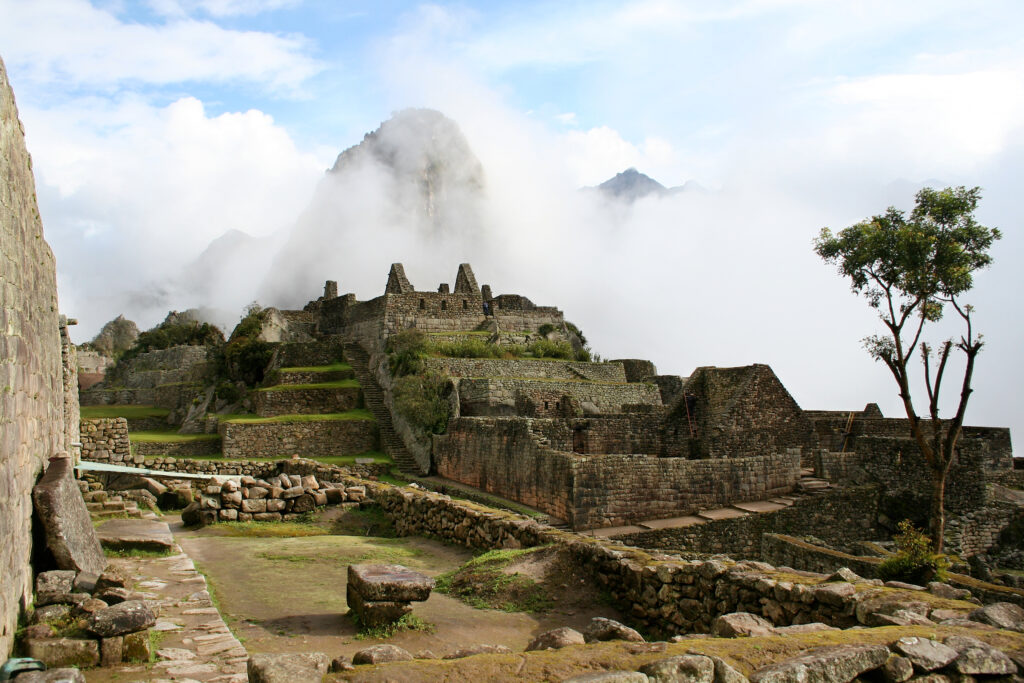A landscape features a grassy complex with a large, tiered structure made of stones that towers upward toward a blue, cloud-filled sky. Mountains taller than the structure are barely visible behind clouds in the distance.