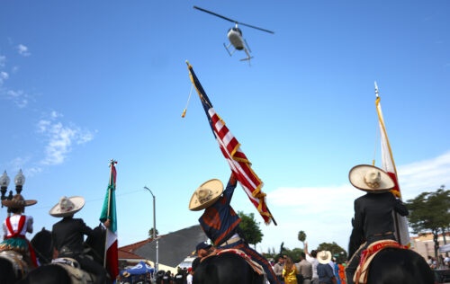 In the foreground, four people wear sombreros and ride on horseback. Three of them carry U.S. and Mexican flags while looking up and gesturing toward a helicopter overhead. A crowd of people walk in front of them.
