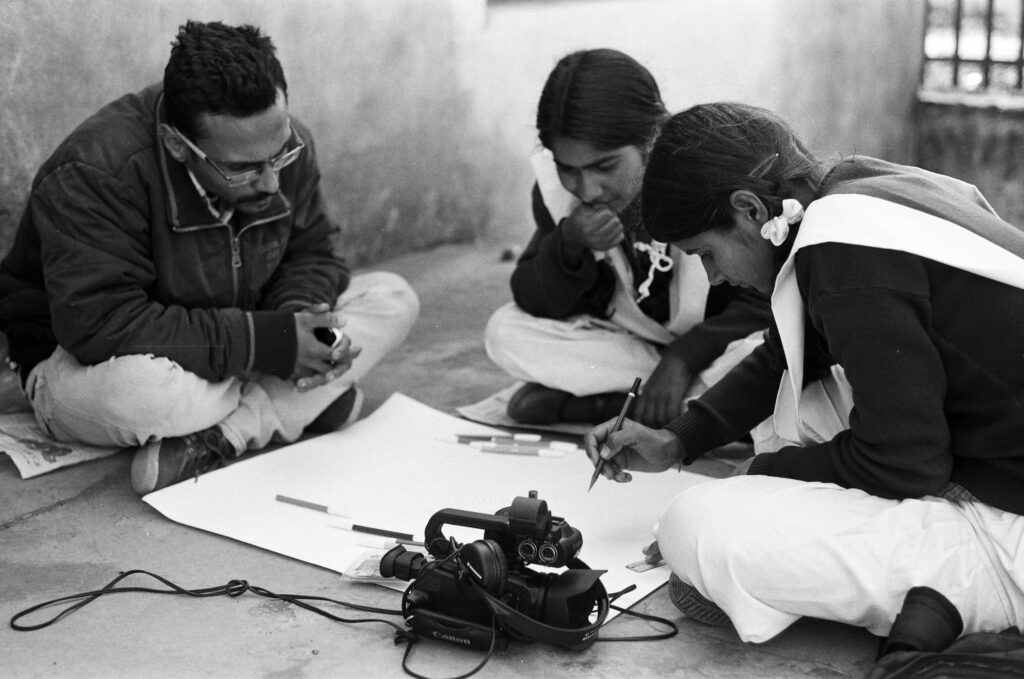 An adult with short hair, a mustache, and glasses, and two young people with long hair held by barrettes—one holding a pen—sit on the floor around a large piece of construction paper, markers, and a camera.
