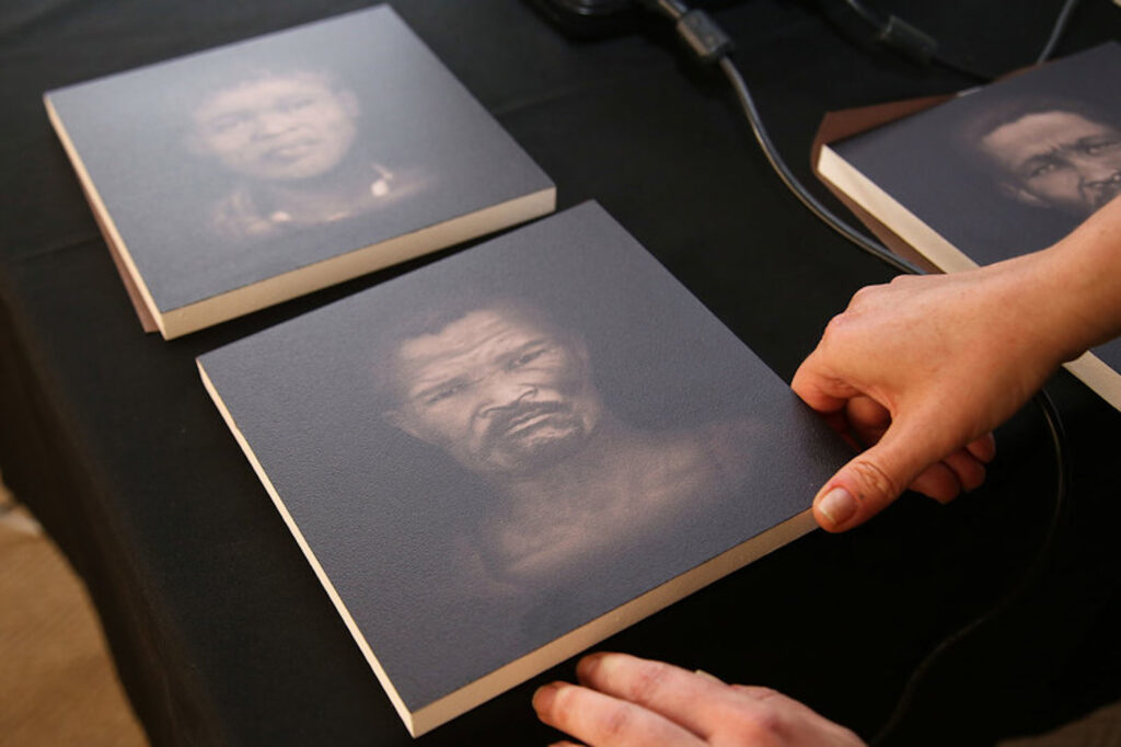 A pair of hands arrange two placards covered with images of two different people’s faces on a table covered with a black tablecloth.