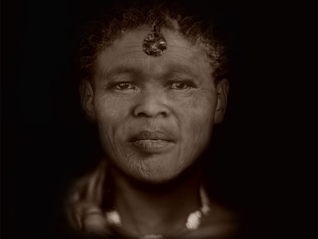 From the shoulders up, a person with short, coiled, graying hair wearing head and neck jewelry stares at the viewer in front of a pitch-black background.