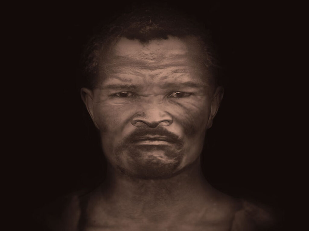 From the shoulders up, a person with short, coiled, black hair and a mustache stares at the viewer in front of a pitch-black background.