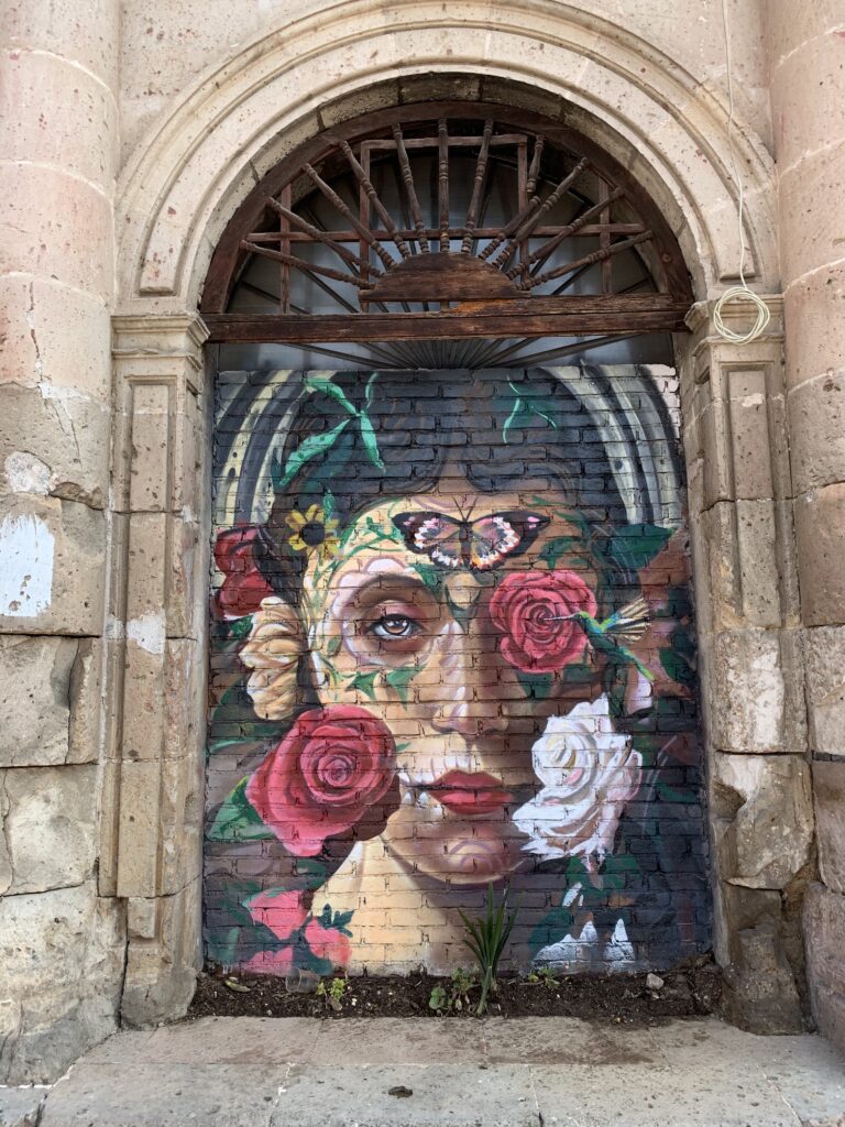 Beneath a stone arch, a large painting covering a brick wall depicts the face of a person wearing red lipstick with roses, other flowers, green leaves, a butterfly, and a hummingbird scattered across it.