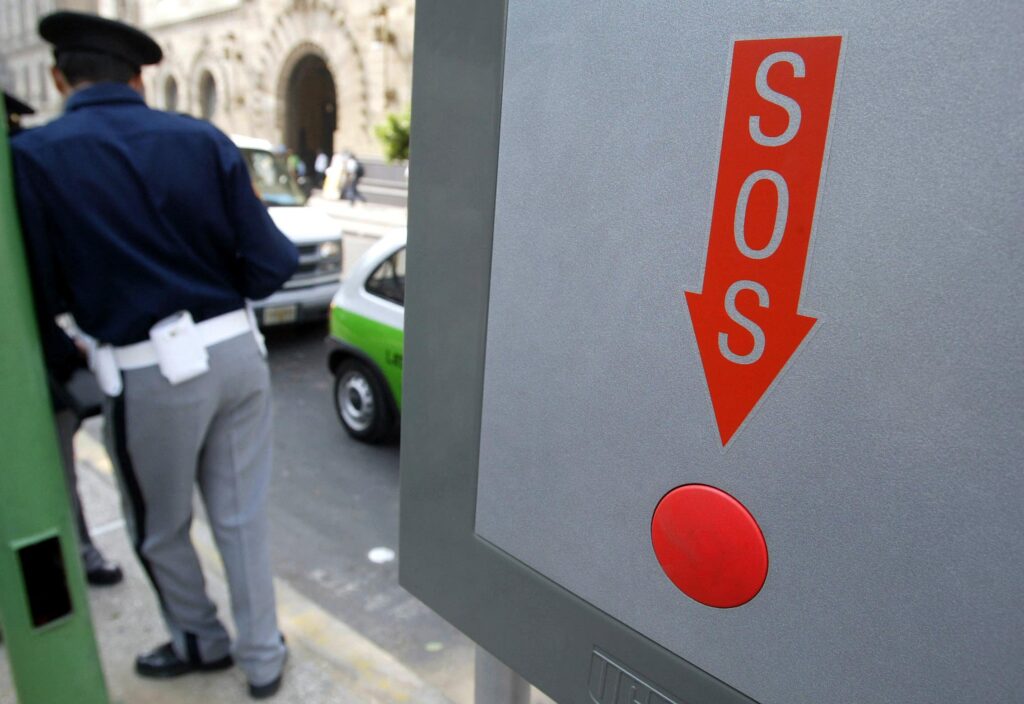 A police officer stands next to a gray sign with “SOS” written inside a red arrow pointing to a red button.