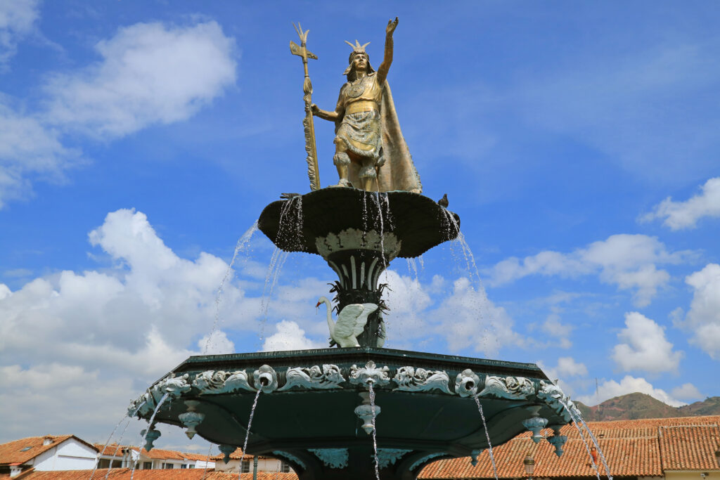 Against a blue sky with white clouds, a golden statue of a person with a staff in their right hand and raising up their left hand sits atop a two-tiered fountain in a public square.