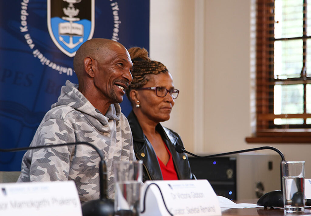 Two people sit at a long table behind white folded name labels and microphones. The left-most person wears a gray camo hoodie and has a mustache and beard, and the other wears earrings, glasses, a red shirt, and a black jacket.