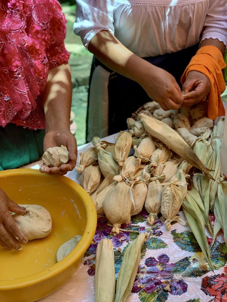 Two people’s hands handle food on a table covered in a floral tablecloth. The hands on the left knead a ball of dough while those on the right use corn husks to package small objects into bundles.