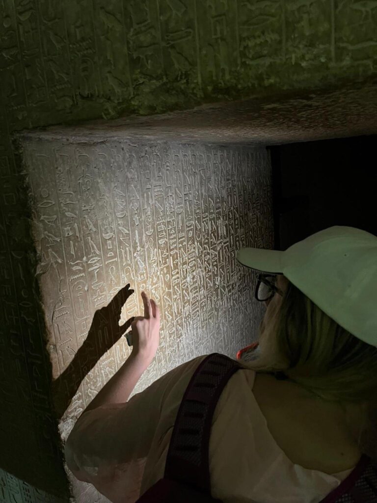 A person wearing a white cap, white shirt, and backpack points at etchings covering a large stone wall in a dark room with a low ceiling.