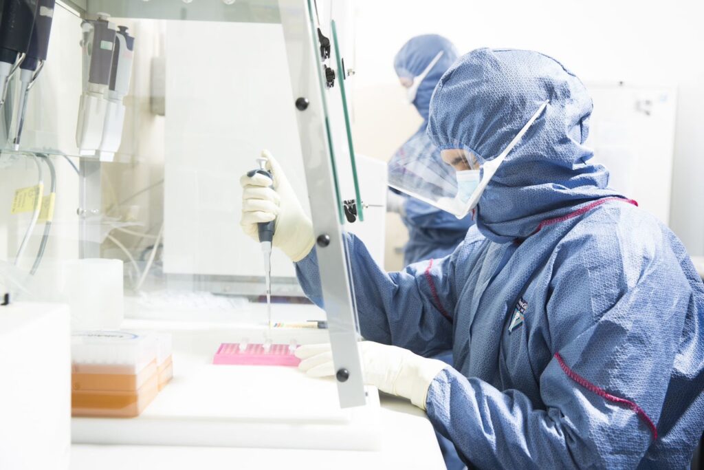 Two people wearing blue full-body hazmat suits, goggles, and white gloves are in a white lab. The person in the foreground sits at a table holding a sample tray on the table with their left hand and a pipette in their right.