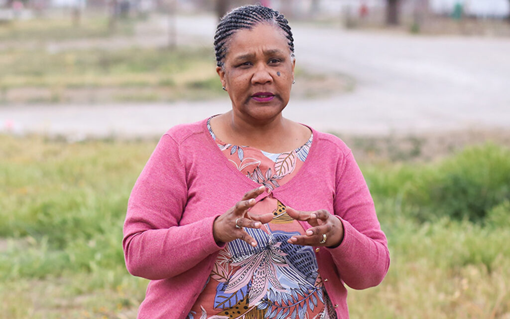 On a grassy field with a road winding in the distance, a person with cornrows wearing fuchsia lipstick, a floral shirt, and a pink cardigan speaks while gesturing with their hands.