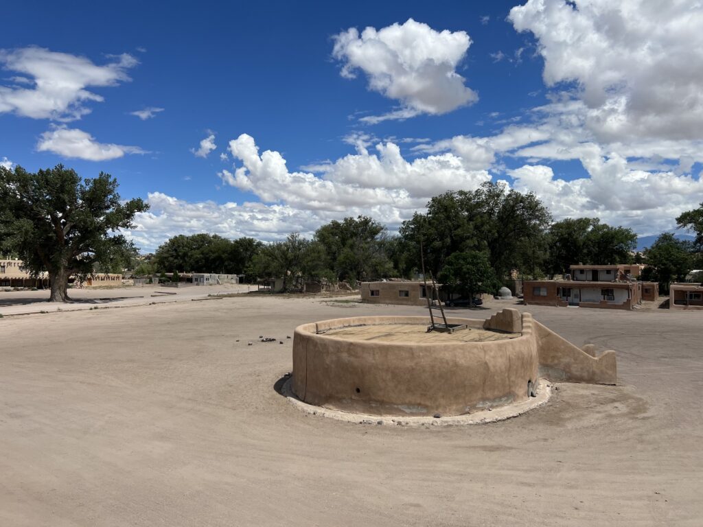 A circular stone structure with a small entry ladder down into it sits in the middle of a flattened dirt area with rectangular buildings, green trees, and a blue sky lining the background.