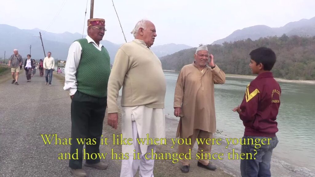 A film still features a child in a maroon jacket talking to three elders near the shore of a river as a group of people walk on the road behind them. Mountains covered in forests line the horizon.