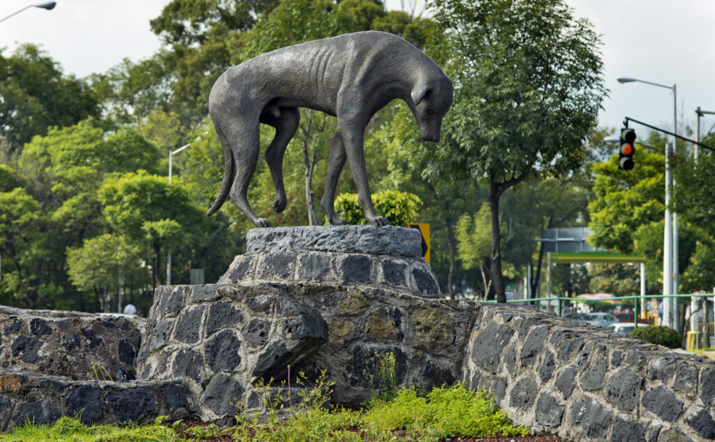 A large metal statue depicting a standing dog with its head down and ribs visible is on top of a curved stone wall with trees along the background and a traffic light to the right.