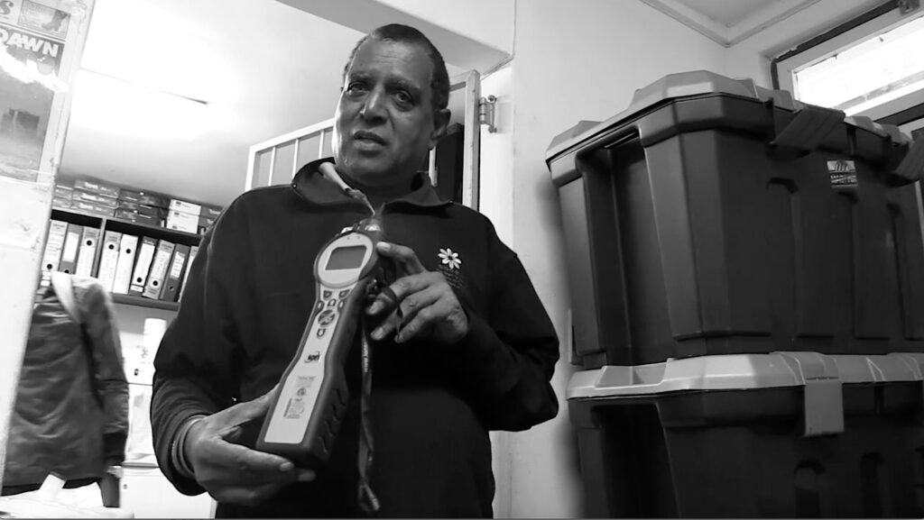 A black-and-white film still features a person with short hair in a sweater next to two large, stacked storage containers. They are pointing their right index finger while holding a tool in their left hand.