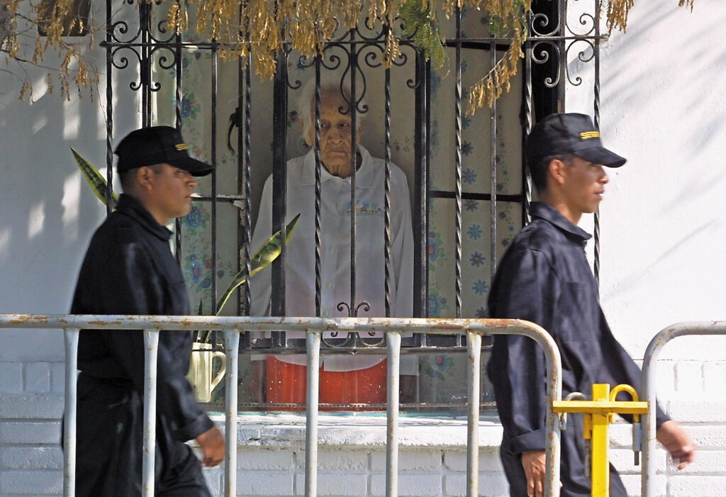 A person with gray hair looks through the barred window of a home at two police officers walking past on a street.