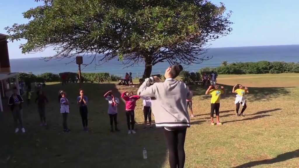 A film still features a person with brown hair up in a bun in a gray sweater and black leggings facing a row of children with their left hand on their shoulder. The children are mimicking this action. Film by student participants, courtesy of Peter Sutoris