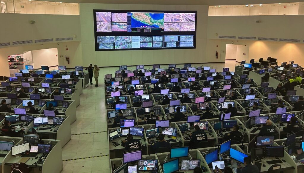 In the center of a huge, multistory room, multiple computers hanging on a wall display images of city streets. In the foreground, dozens of people sit at cubicles looking at similar computer images on their desks.