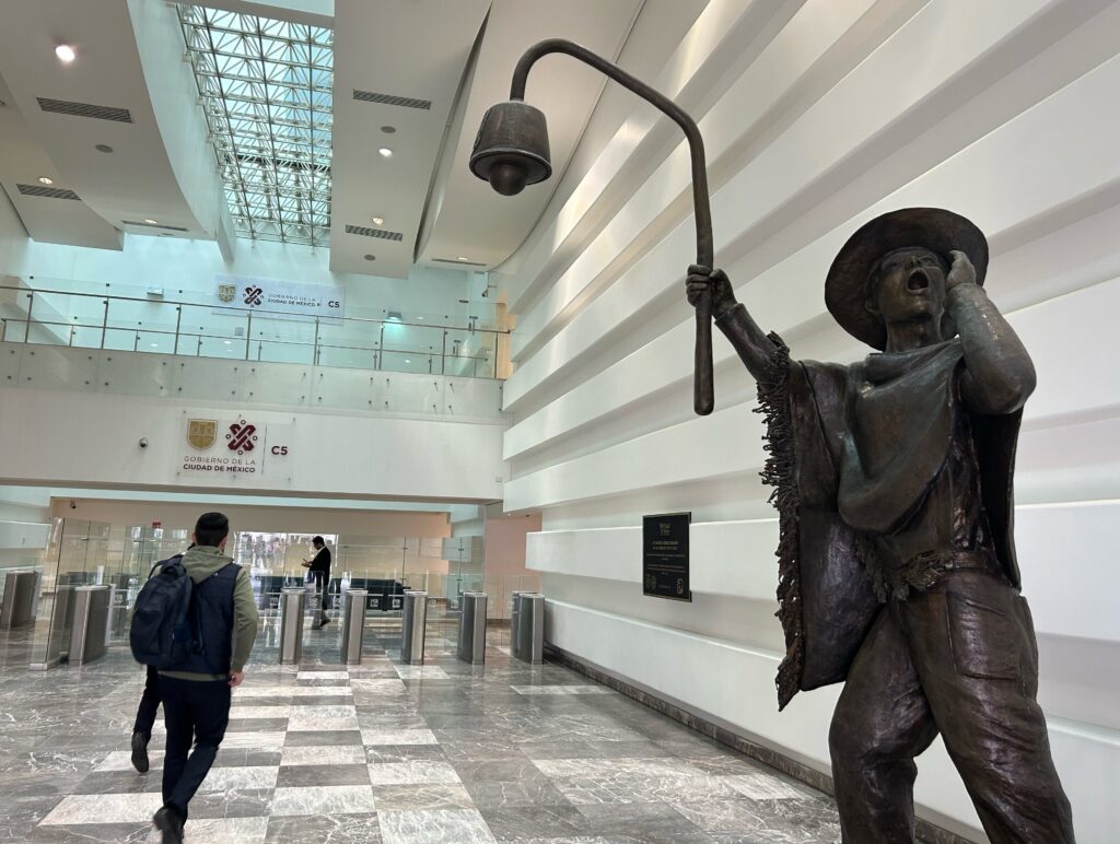 Inside a large white and gray building stands a black statue of a man wearing a hat and poncho. He holds one hand to his open mouth, as if calling out; in the other hand, he carries a pole with a modern security camera on the end of it.
