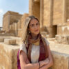 A woman wearing a scarf and earrings stands before ancient buildings made of sandstone.