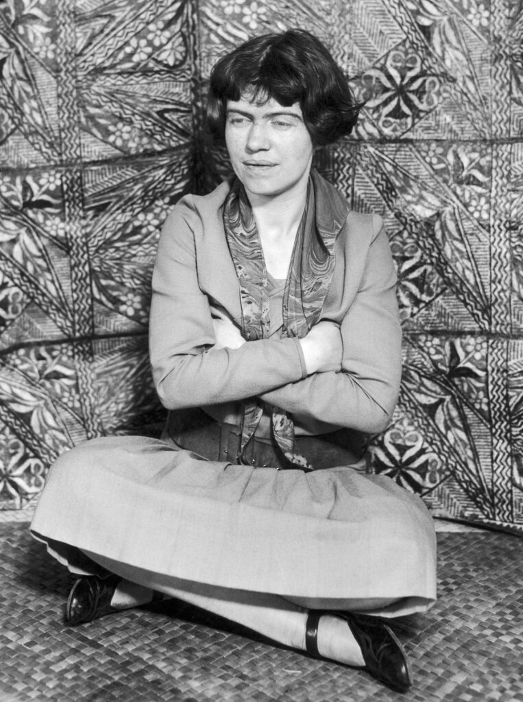 A dark-haired woman sits cross-legged in a dress and black Mary Jane shoes, leaning against a wall imprinted with leaves and other designs.