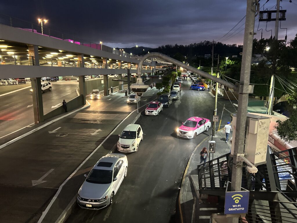 At twilight, multiple cars proceed along a road next to a large parking garage. In the foreground stands a security camera that looks like a dark street light.