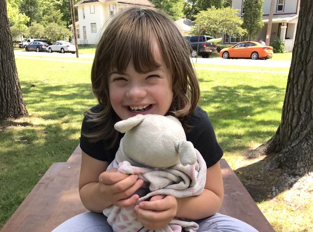 A smiling girl with shoulder-length brown hair in a black T-shirt and gray leggings sits on a wooden table in a grassy park and hugs a stuffed animal wrapped in a pink and white blanket.