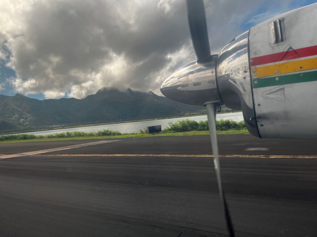 A plane propeller peeks out from the right side of the image, with an island landscape in the background with green horizon, ocean, and cloud covered mountains.