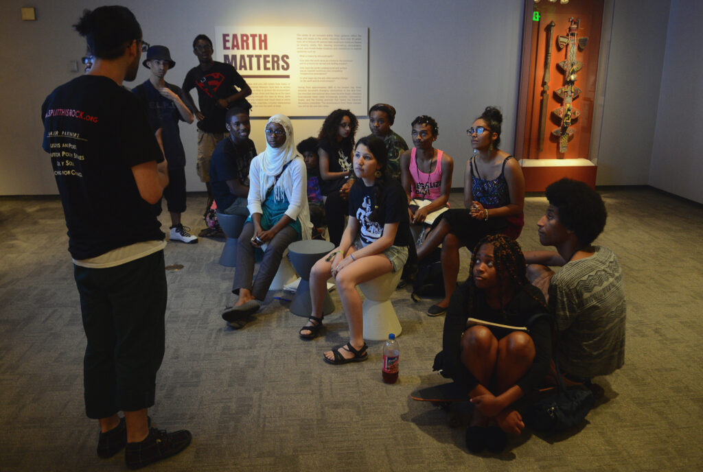 A group of people sit in a carpeted room facing a person who is standing. On the illuminated back wall are a vertical display case holding carved wooden objects and a large placard under a header of capitalized brown letters that reads, “Earth Matters.”