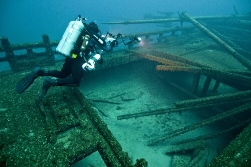 A blue underwater scene features a scuba diver in a black wetsuit with a silver oxygen tank on their back. They point a light at a shipwreck of broken, moss-covered wooden beams.