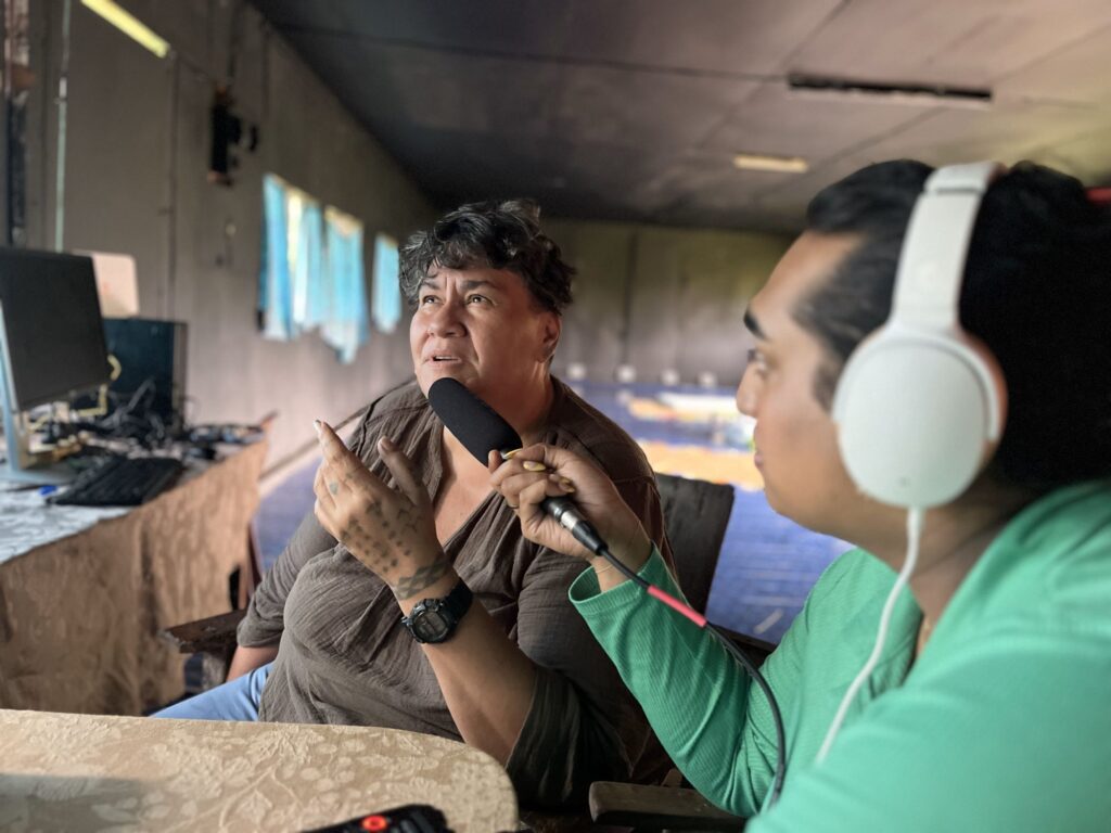 A women in a green sweater and earphones holds a microphone up to another woman with short hair who is gesticulating with a raised hand thoughtfully.